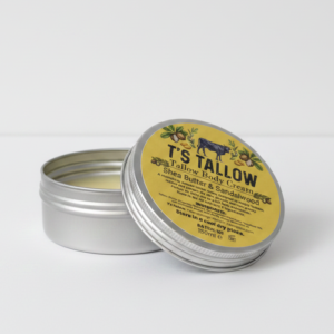 Metal tin of T's Tallow Sheba Butter & Sandalwood body cream, open with the lid resting beside it on a white surface, yellow label visible
