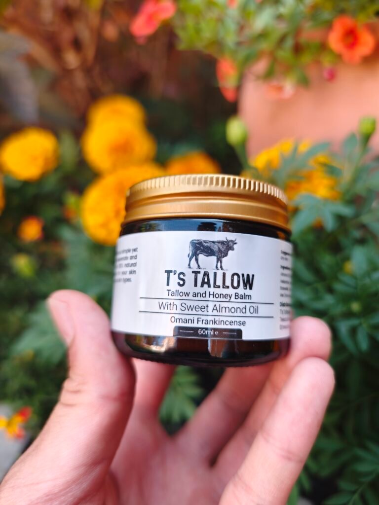 Jar of T's Tallow balm held by hands, with a colorful flower garden blurred in the background.
