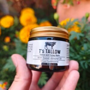 Jar of T's Tallow balm held by hands, with a colorful flower garden blurred in the background.